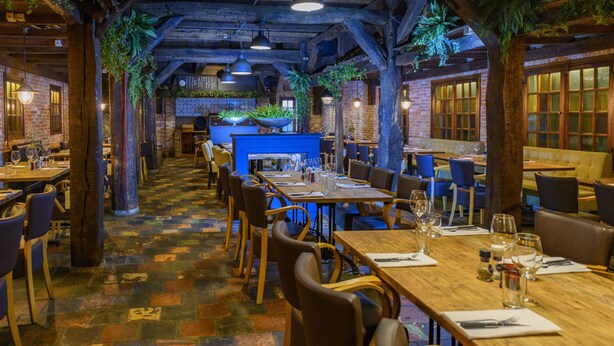 A restaurant with wooden tables and chairs, a brick wall, and a blue counter. - Grand Hotel Amstelveen (Amstelveen)