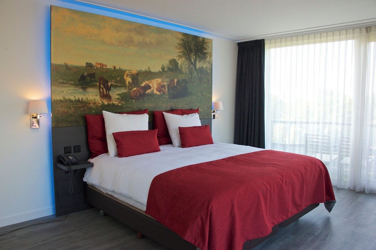 A hotel room with a large painting of a pastoral scene, a bed with red and white bedding, a bedside table with a lamp, and a large window with sheer curtains.