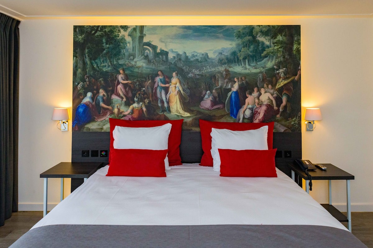 Habitación de hotel con una cama grande, almohadas rojas y blancas, un cuadro en la pared, dos mesitas de noche con lámparas y un teléfono sobre una de ellas.