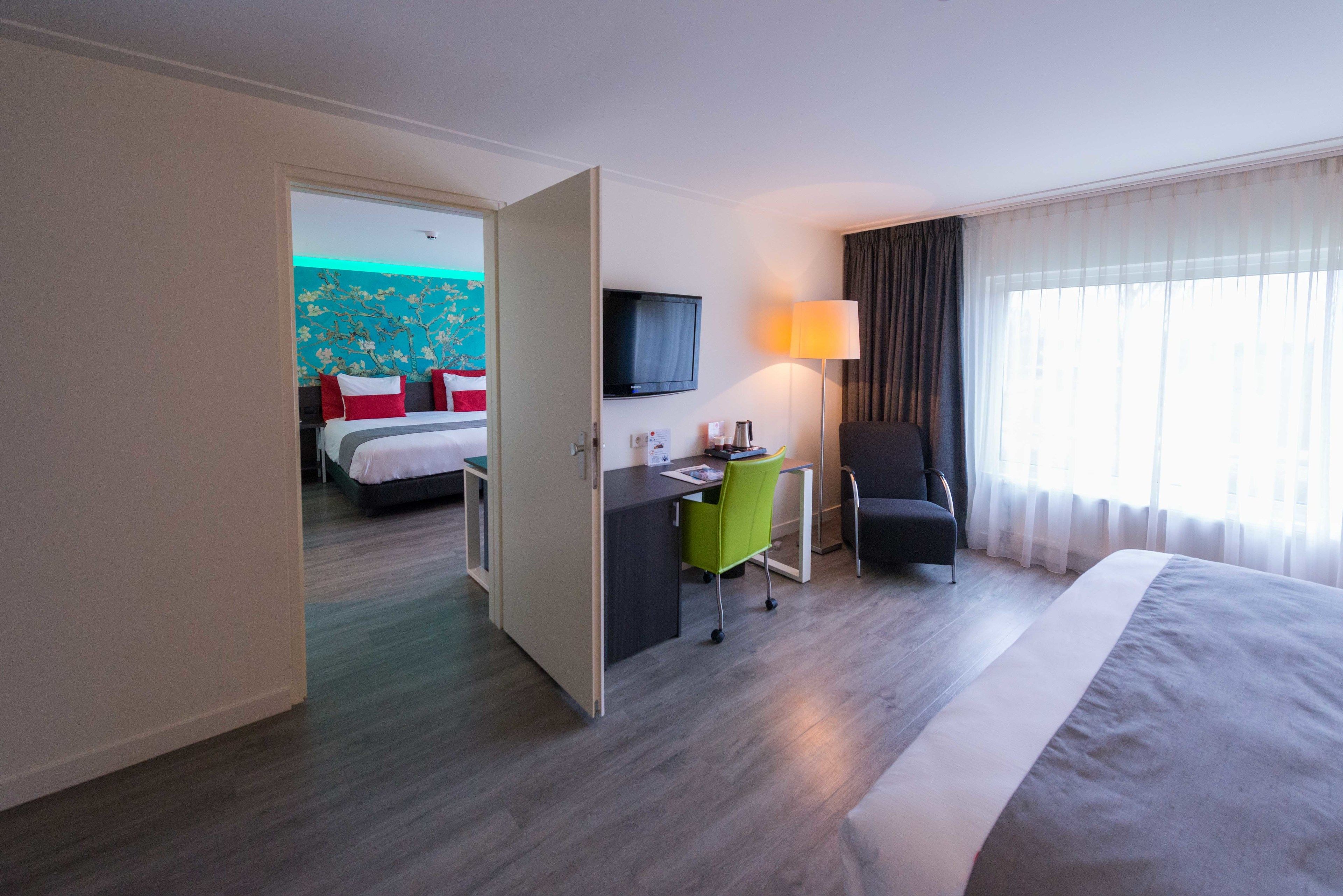 Una habitación de hotel con cama, escritorio, silla, televisor y ventana con cortinas.