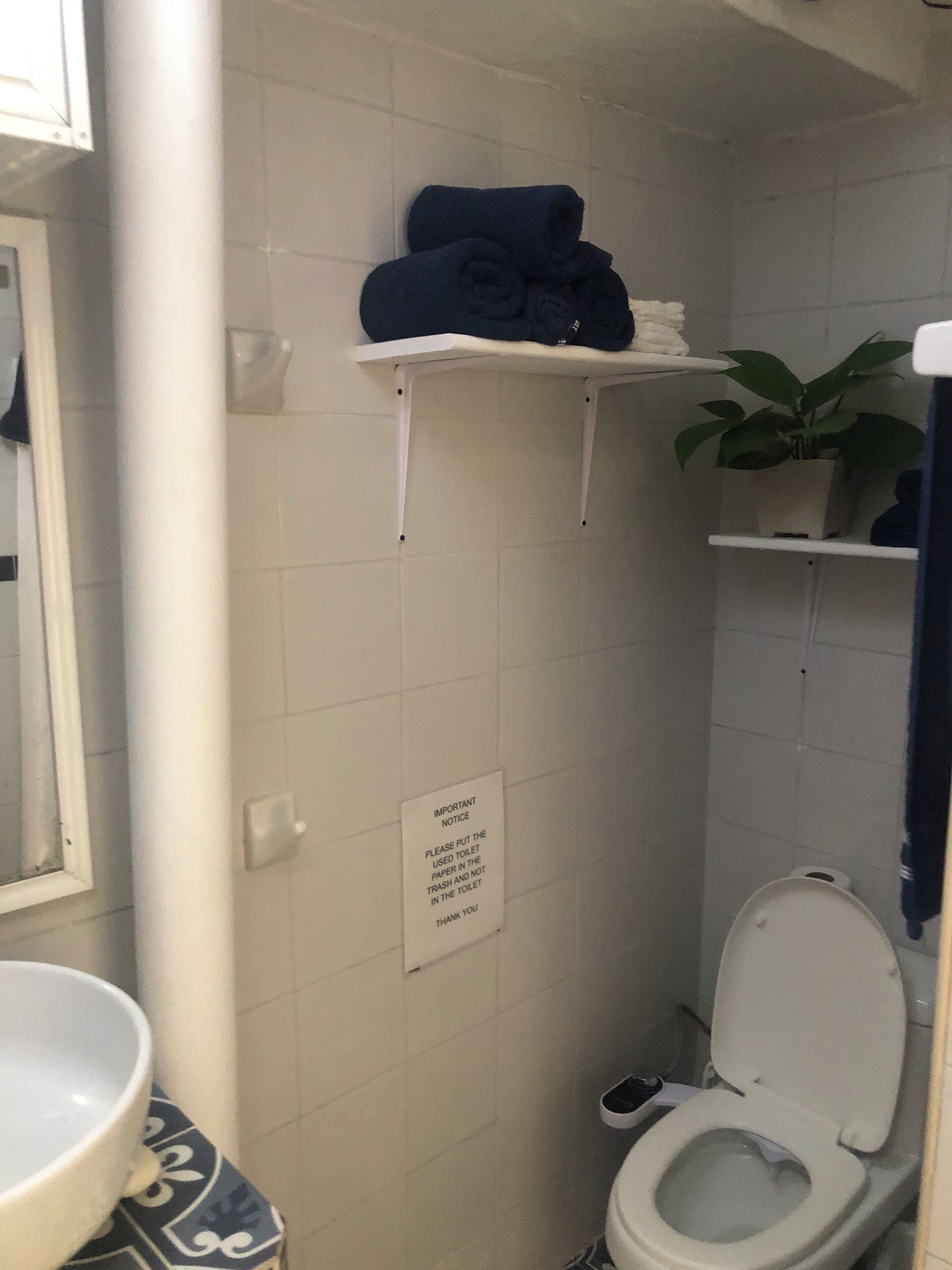 A small bathroom with a toilet, a sink, and a shelf with towels. There is a sign on the wall with instructions for using the toilet.