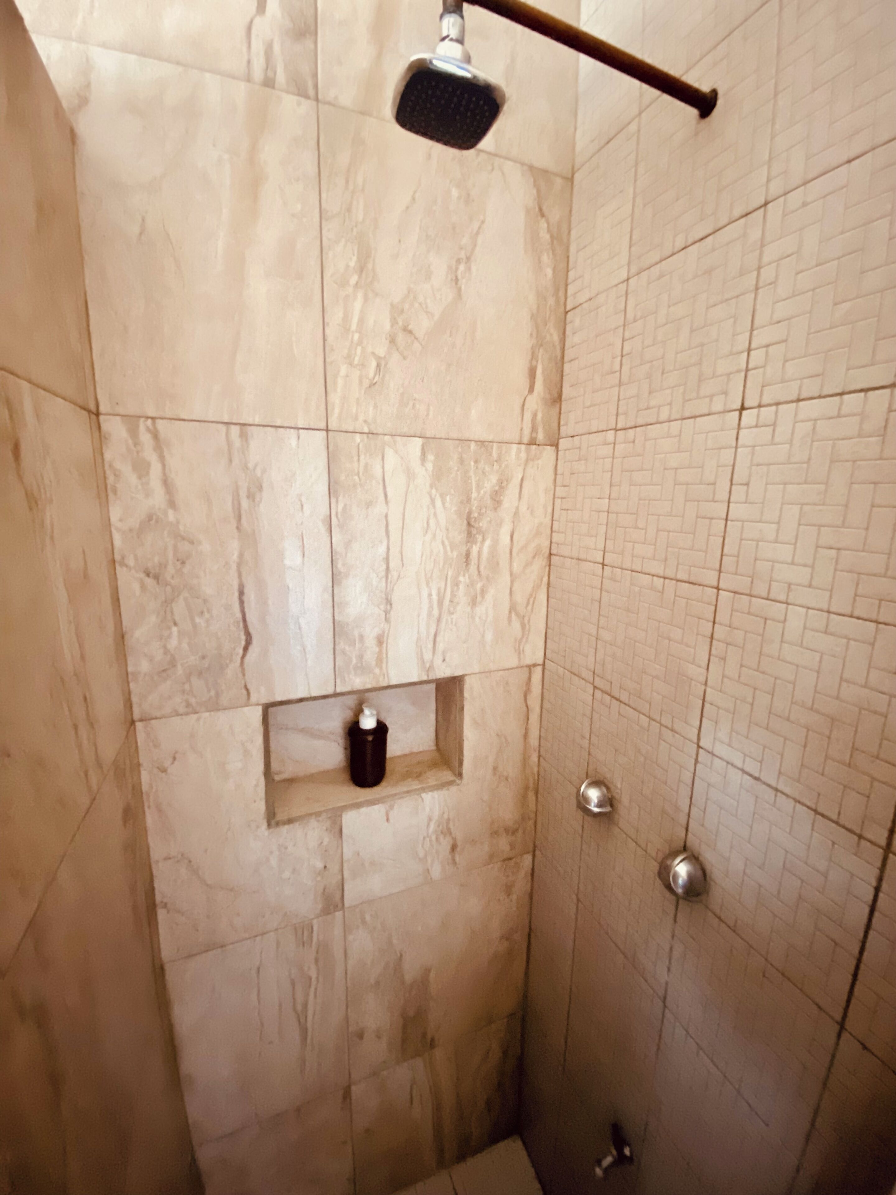 Douchegedeelte met een handdouche, een plankje met een fles en een betegelde muur.