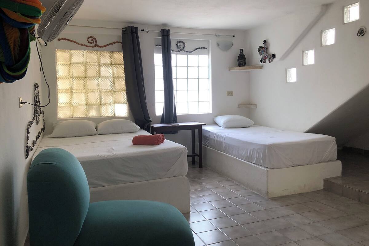 Un dormitorio con dos camas, una mesita, una silla, un ventilador y una ventana con cortinas.
