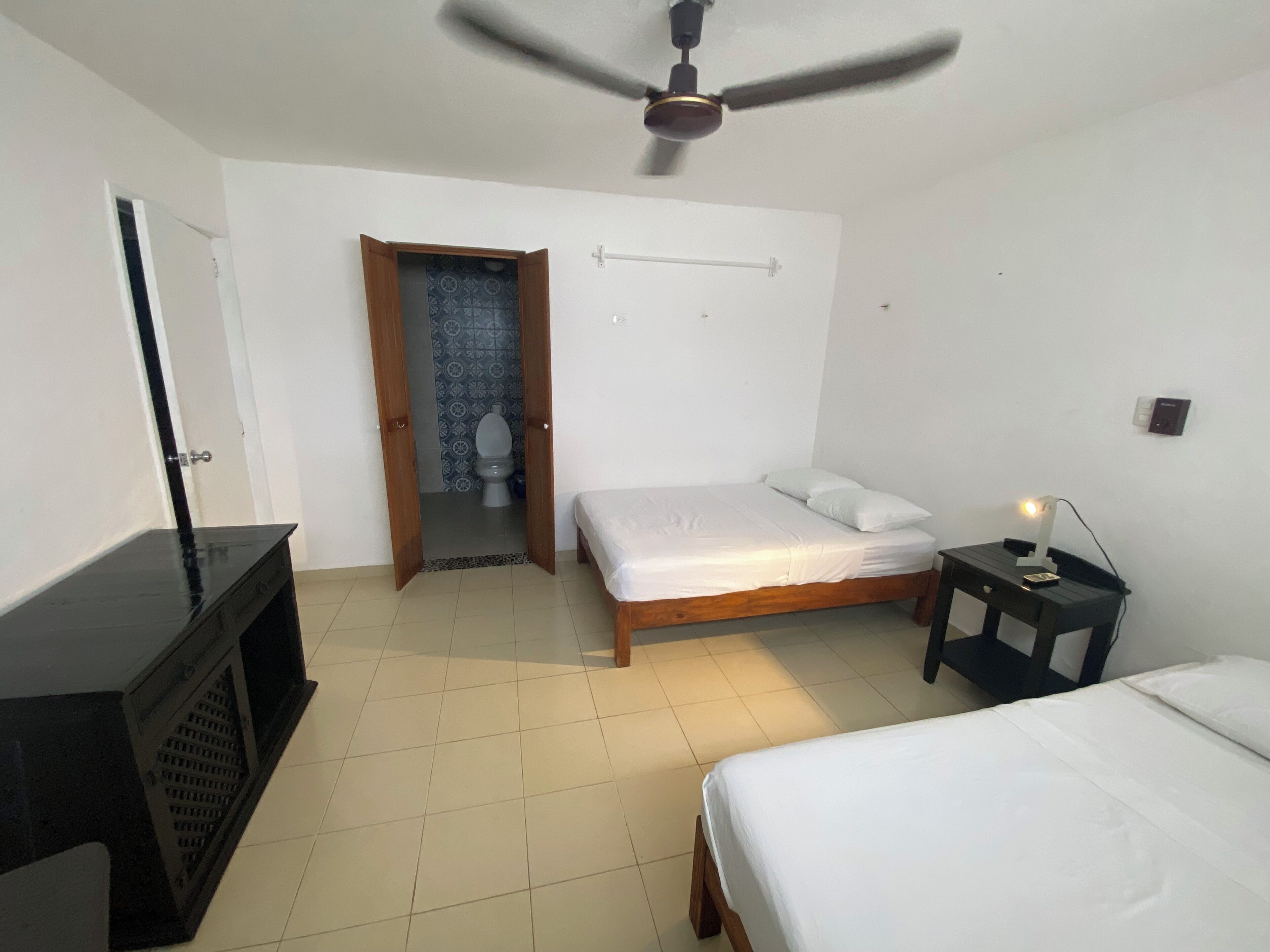Una camera da letto con un letto, un comodino con lampada, una ventola a soffitto, un armadio e un bagno visibile attraverso una porta aperta.