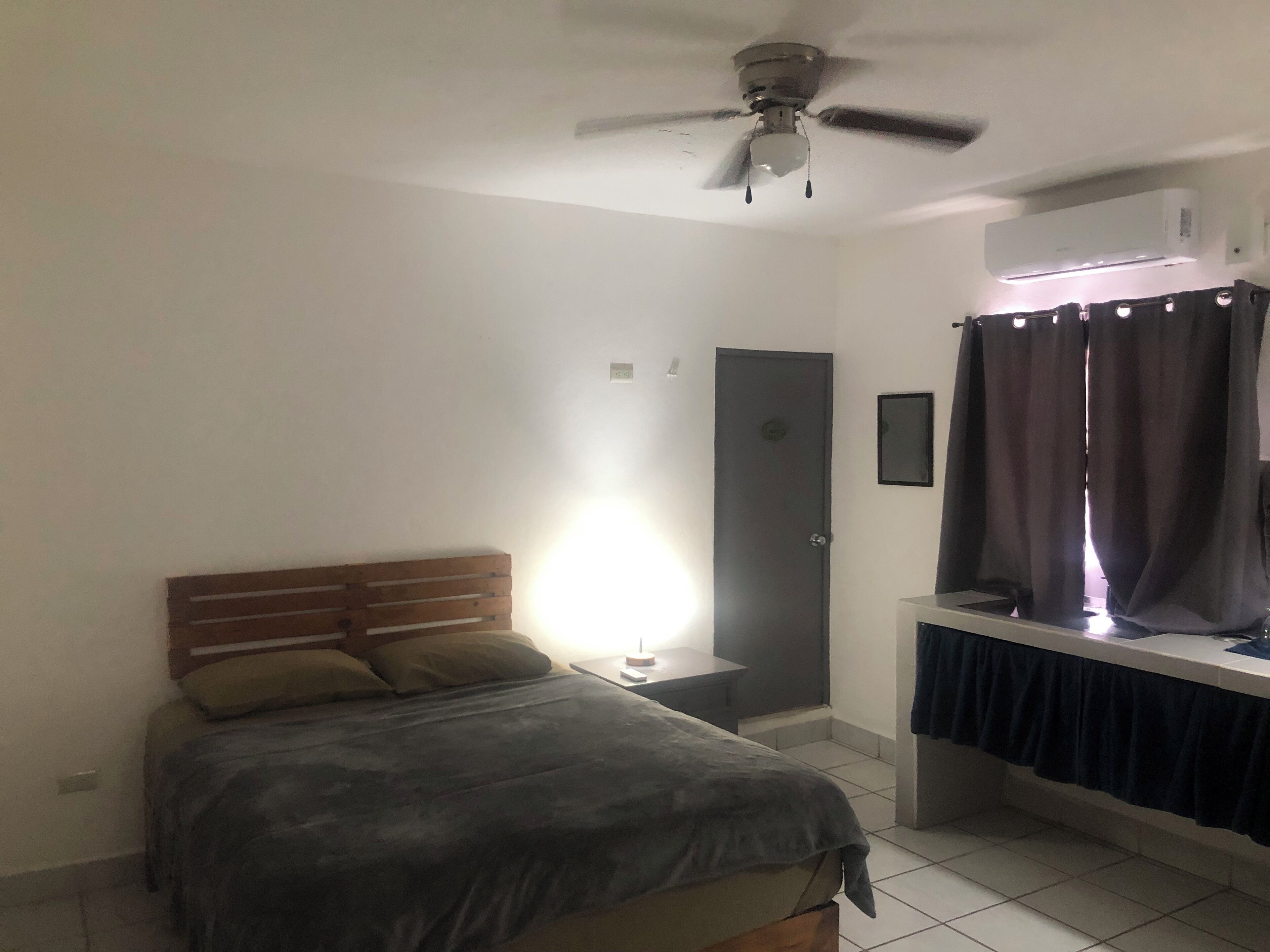 Ein Schlafzimmer mit Bett, Deckenventilator, Klimaanlage, Nachttisch und einem Fenster mit Vorhängen.
