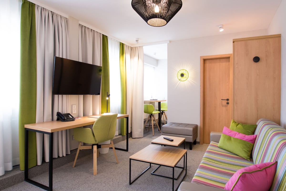 Ein modernes Hotelzimmer mit Schreibtisch aus Holz, grünem Sessel, Sofa und Fernseher.