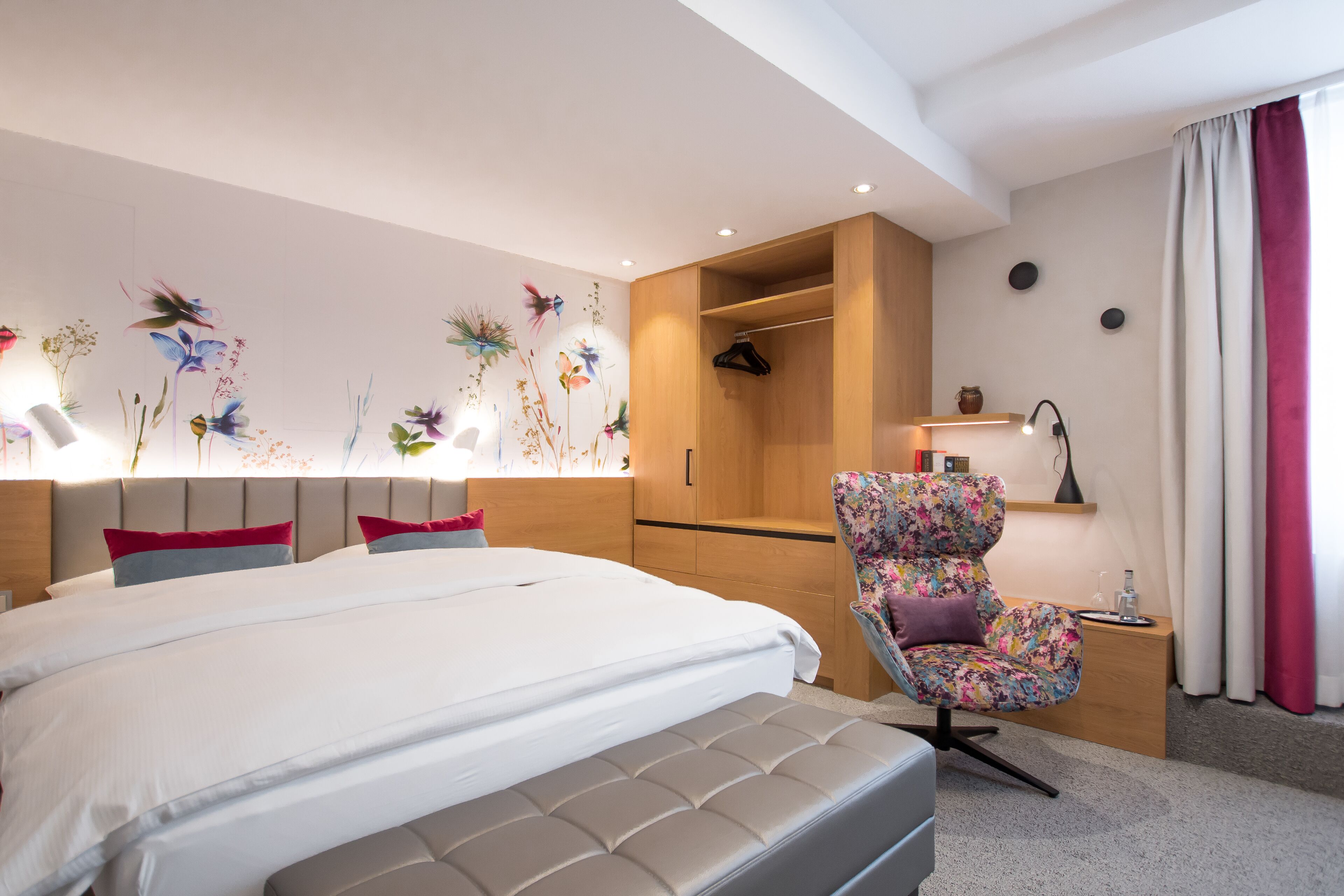 Una habitación de hotel moderna con una cama grande, una zona de estar con un sillón floral, un armario de madera y una pared decorada con motivos florales.
