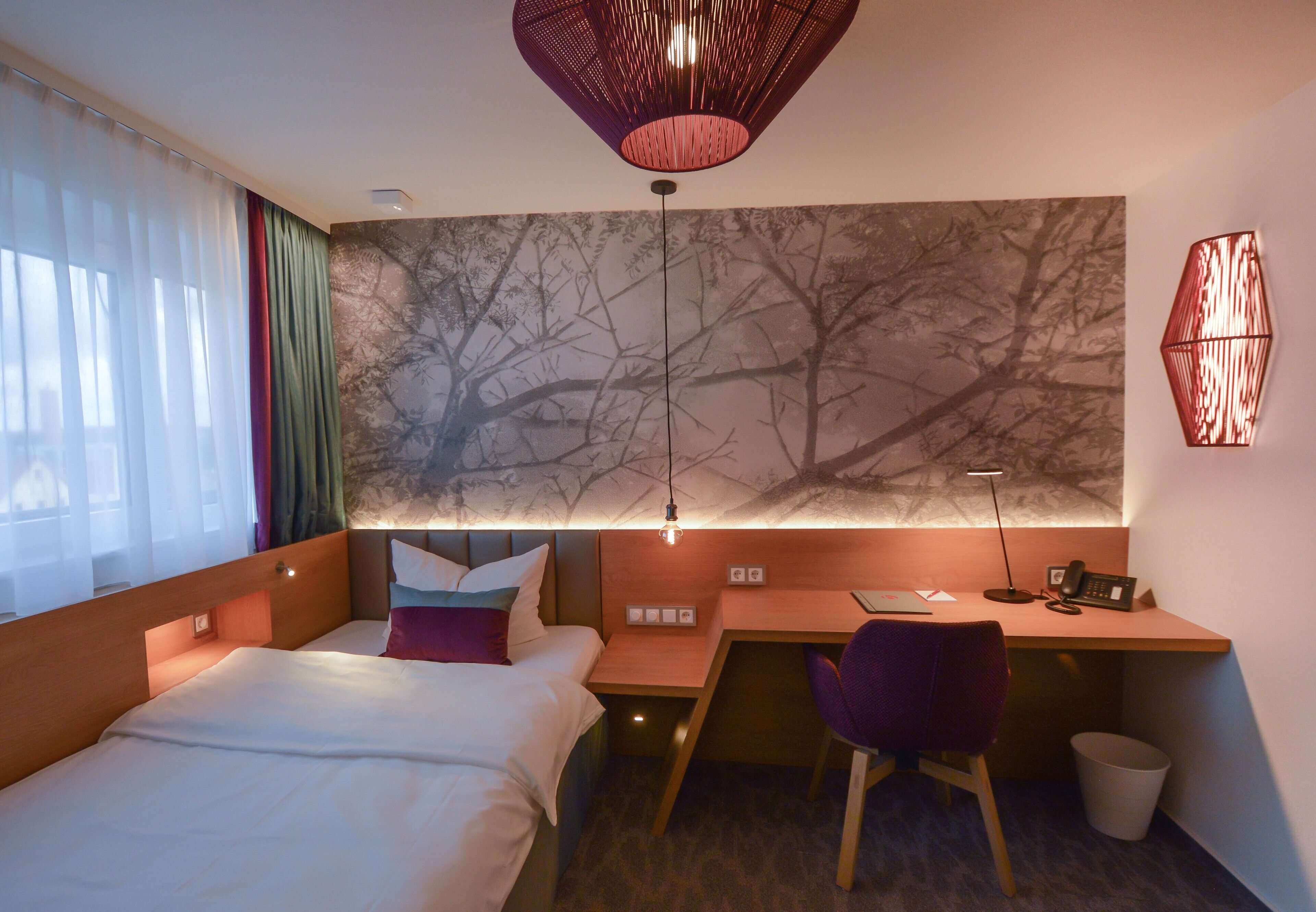 A modern hotel room with a bed, desk, chair, and a large tree mural on the wall.