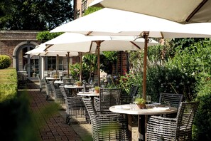 An outdoor patio with wicker tables and chairs, white umbrellas, and a brick building in the background. - Great Fosters - A Small Luxury Hotel (Egham)