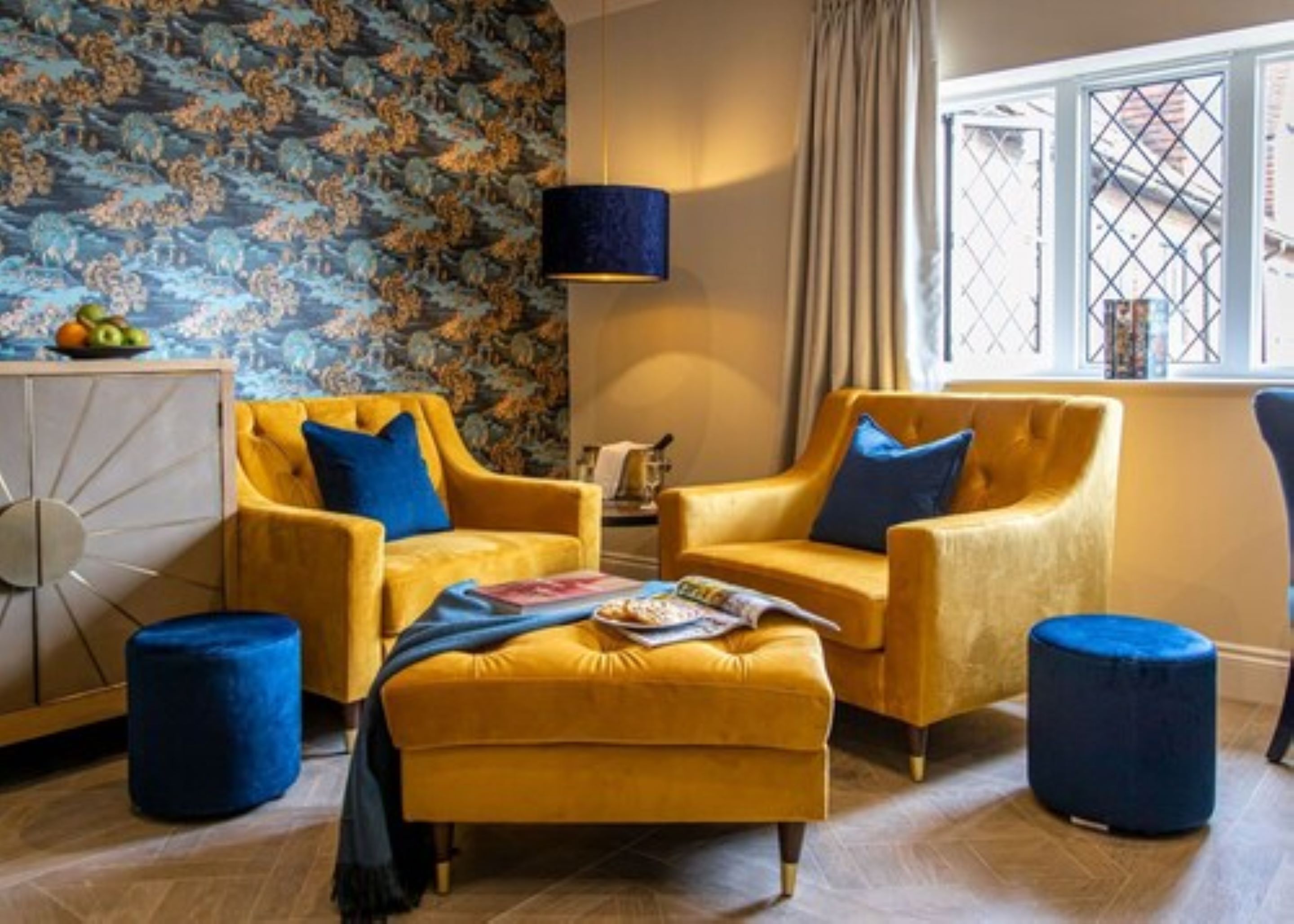 A room with two yellow armchairs, a matching ottoman, and two blue ottomans. A large window with curtains is visible, and a wall with a floral pattern is present.
