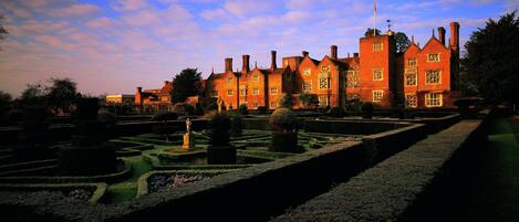 A large, historic building with multiple chimneys and a well-manicured garden with geometric hedges and a statue.