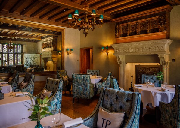 Comedor con vigas de madera, una araña y decoraciones murales elaboradas. Las mesas están dispuestas con manteles blancos y sillas tapizadas en tela azul.