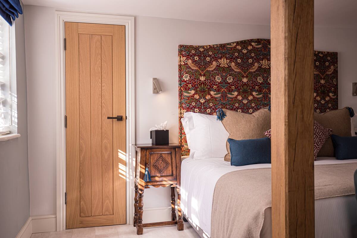 Un dormitorio con una puerta de madera, una mesita de noche con una caja de pañuelos, una cama con varios almohadones y un tapiz decorativo en la pared.