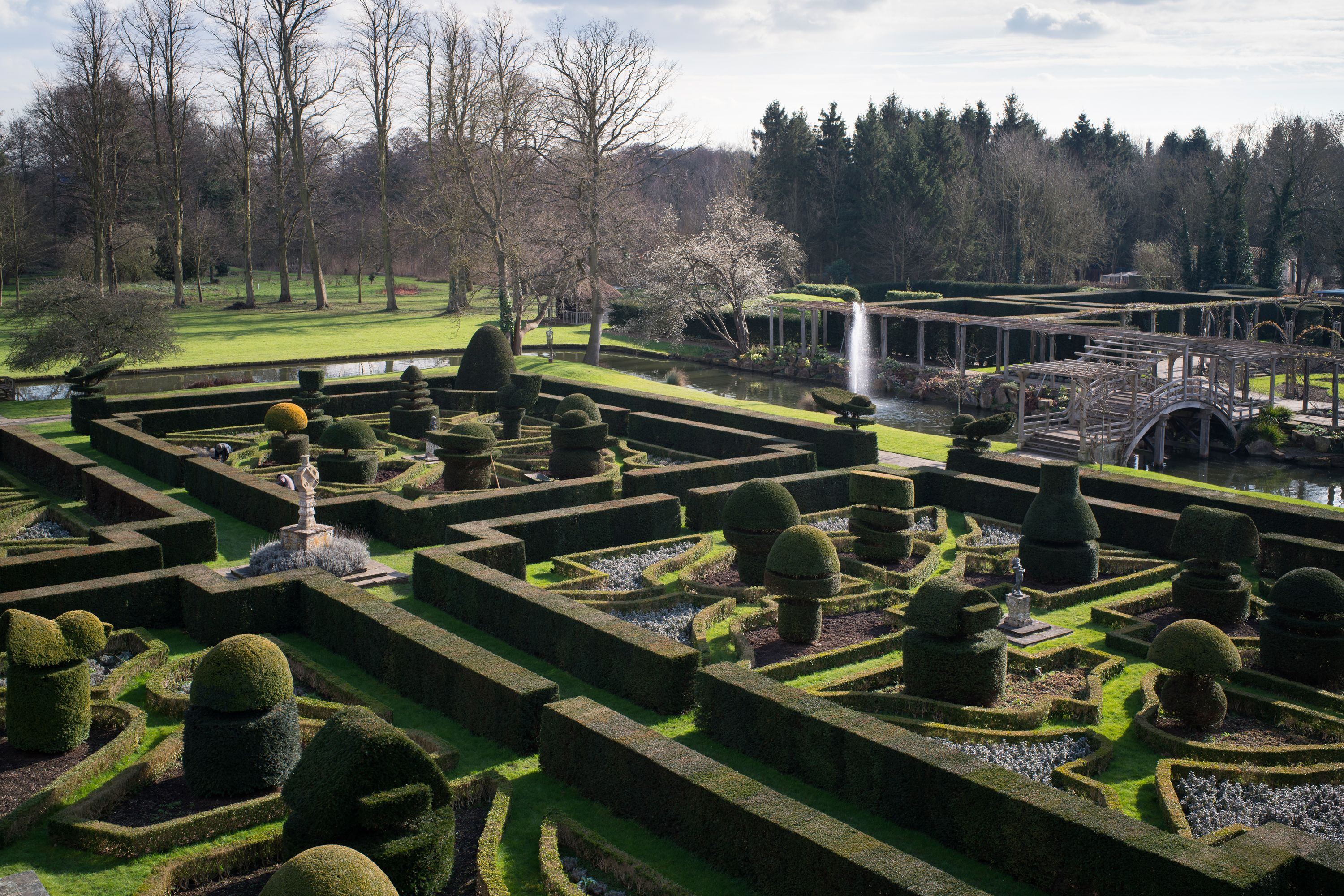A formal garden with geometric hedges, a central fountain, and a bridge over a water feature.