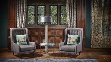 Two armchairs with decorative cushions, a tall floor lamp, a small round table with a glass top, and a patterned tapestry on the wall.