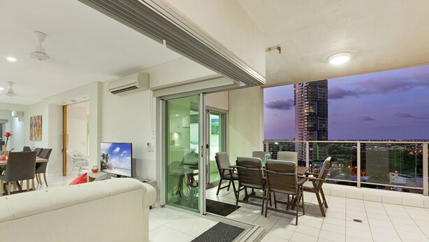 A modern living room with a sofa, dining table, and chairs, a TV, and a balcony with a city view. - Beachlife Holiday Apartments (Darwin)