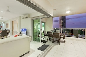A modern living room with a sofa, dining table, and chairs, a TV, and a balcony with a city view. - Beachlife Holiday Apartments (Darwin)