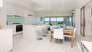 A modern kitchen with white cabinets, a central island with bar stools, and a dining area with a wooden table and white chairs. - Beachlife Holiday Apartments (Darwin)