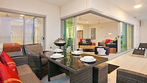 A modern living room with a glass door leading to a balcony, wicker furniture, and a dining table set for four. - Beachlife Holiday Apartments (Darwin)