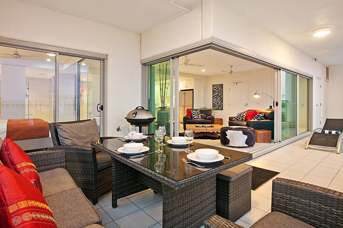 Una sala de estar moderna con un ventanal que da a un balcón, muebles de mimbre y una mesa de comedor para cuatro.