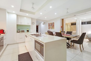 A modern kitchen with white cabinets, a central island, and a dining area with chairs and a table. - Beachlife Holiday Apartments (Darwin)