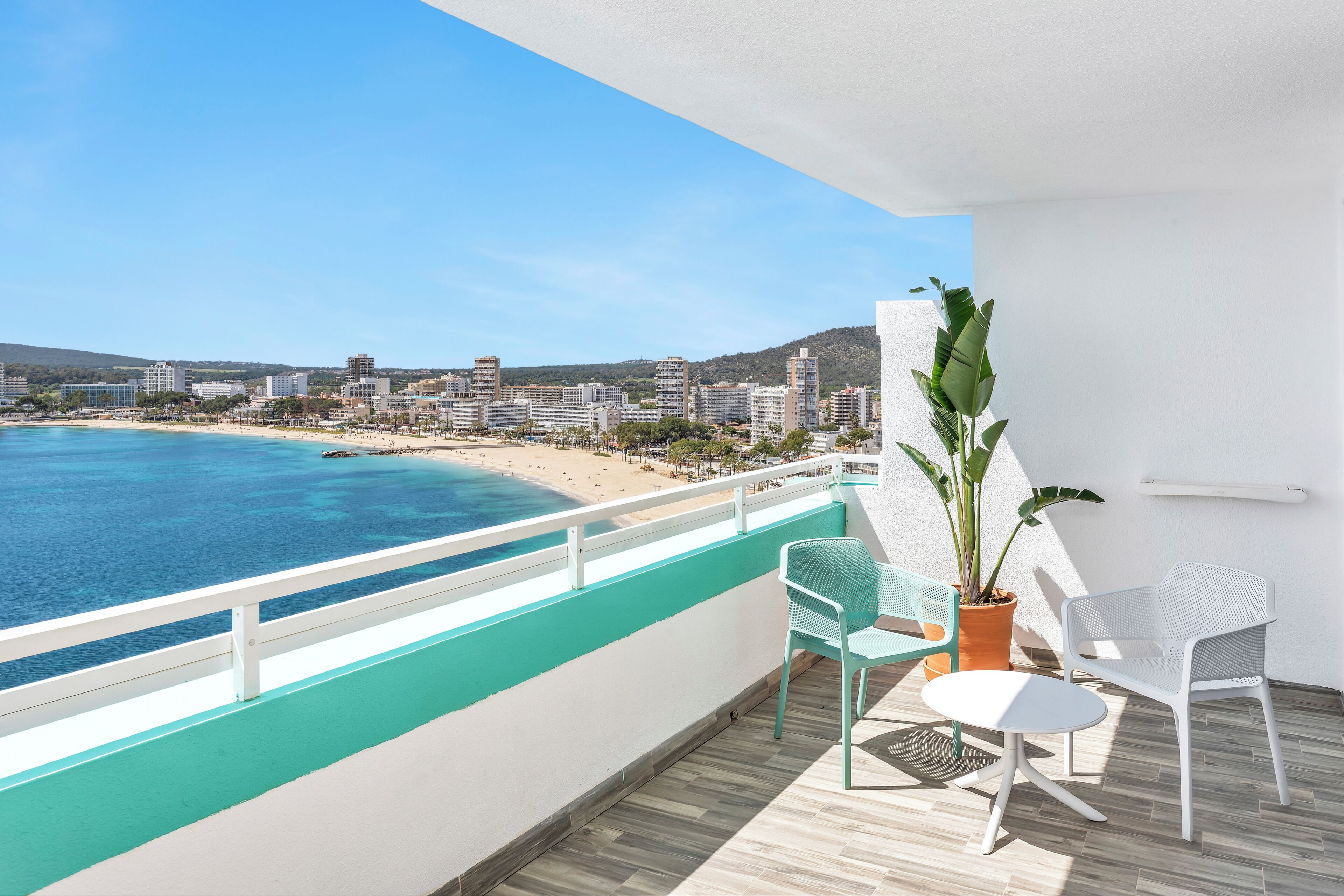 A balcony with a view of a beach and cityscape, including a potted plant and two chairs.