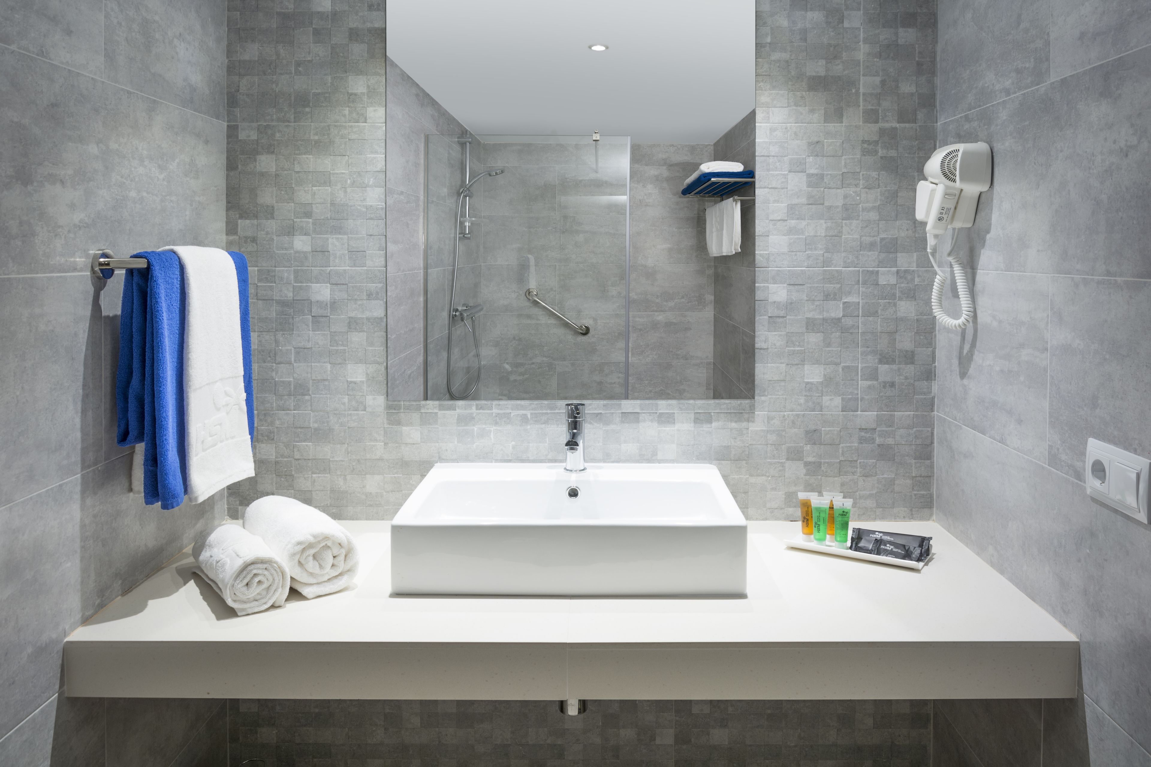 A modern bathroom with a white sink, two towels, a handheld showerhead, and a hairdryer mounted on the wall.