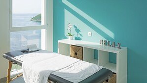Une salle de massage avec vue sur la mer, une table de massage blanche, une console blanche agrémentée d’une plante et un panneau indiquant « RELAX ».