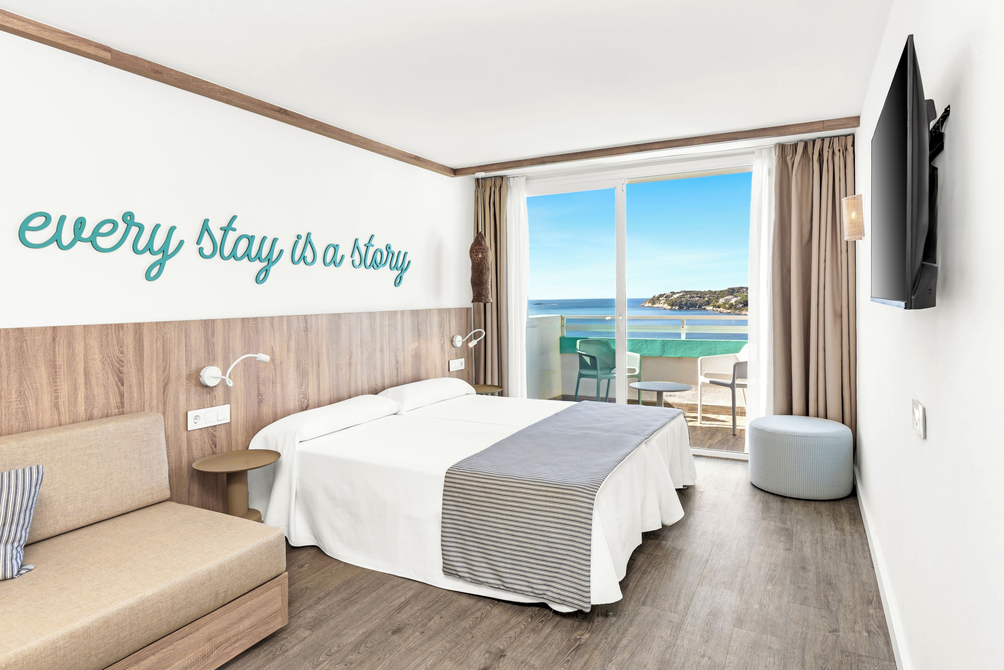 A hotel room with a bed, a sofa, a TV, a balcony with a view of the sea, and a sign on the wall.