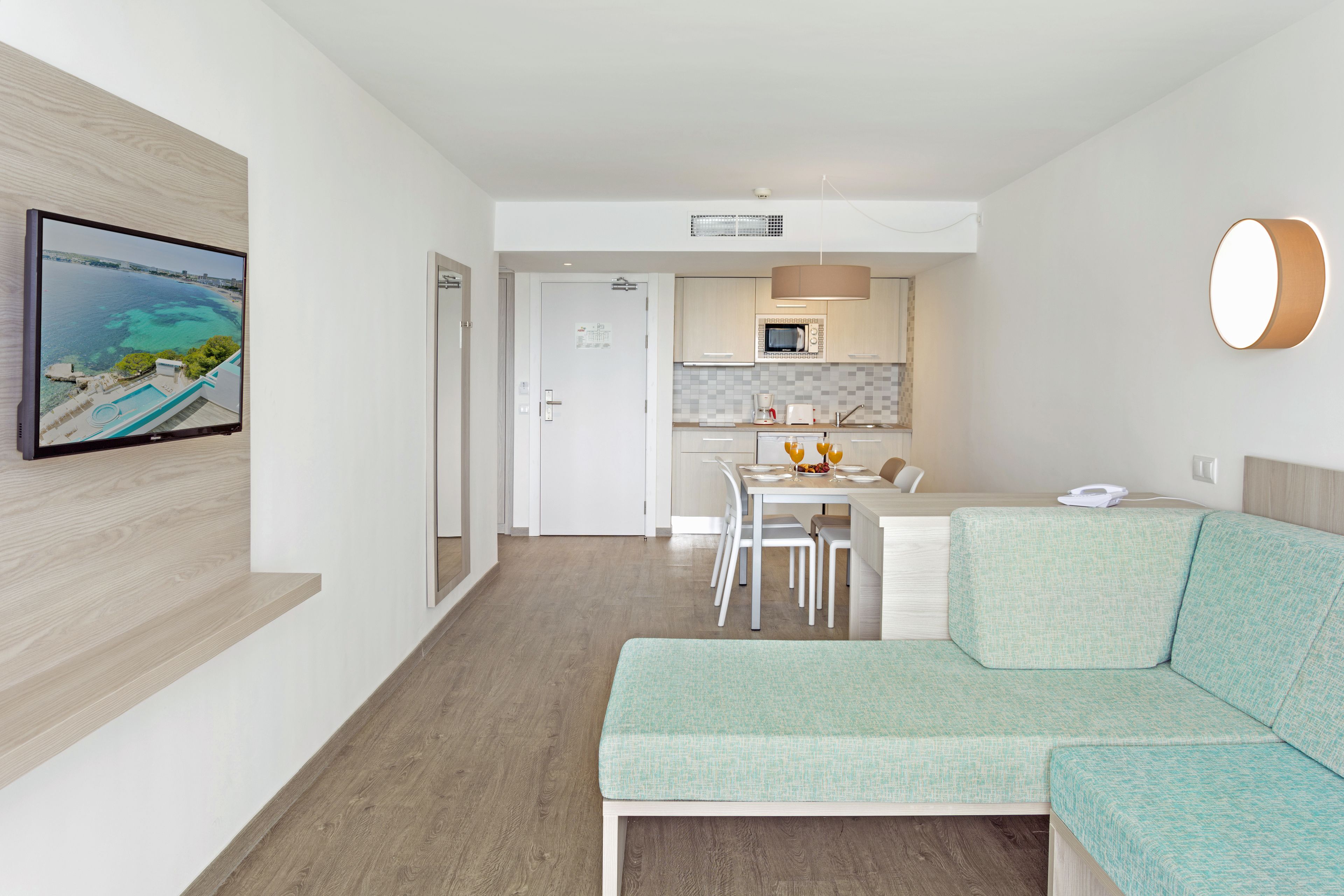 Una sala de estar moderna con un sofá verde claro, un televisor de pantalla plana montado en la pared, una zona de comedor con una mesa y sillas blancas, y una cocina con armarios blancos y un microondas.