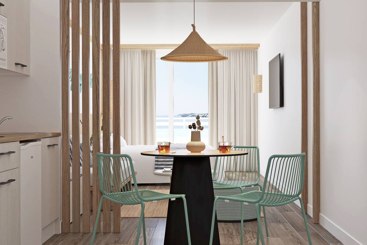 A dining area with a round table, four green chairs, a woven pendant light, and a view of the sea through large windows.
