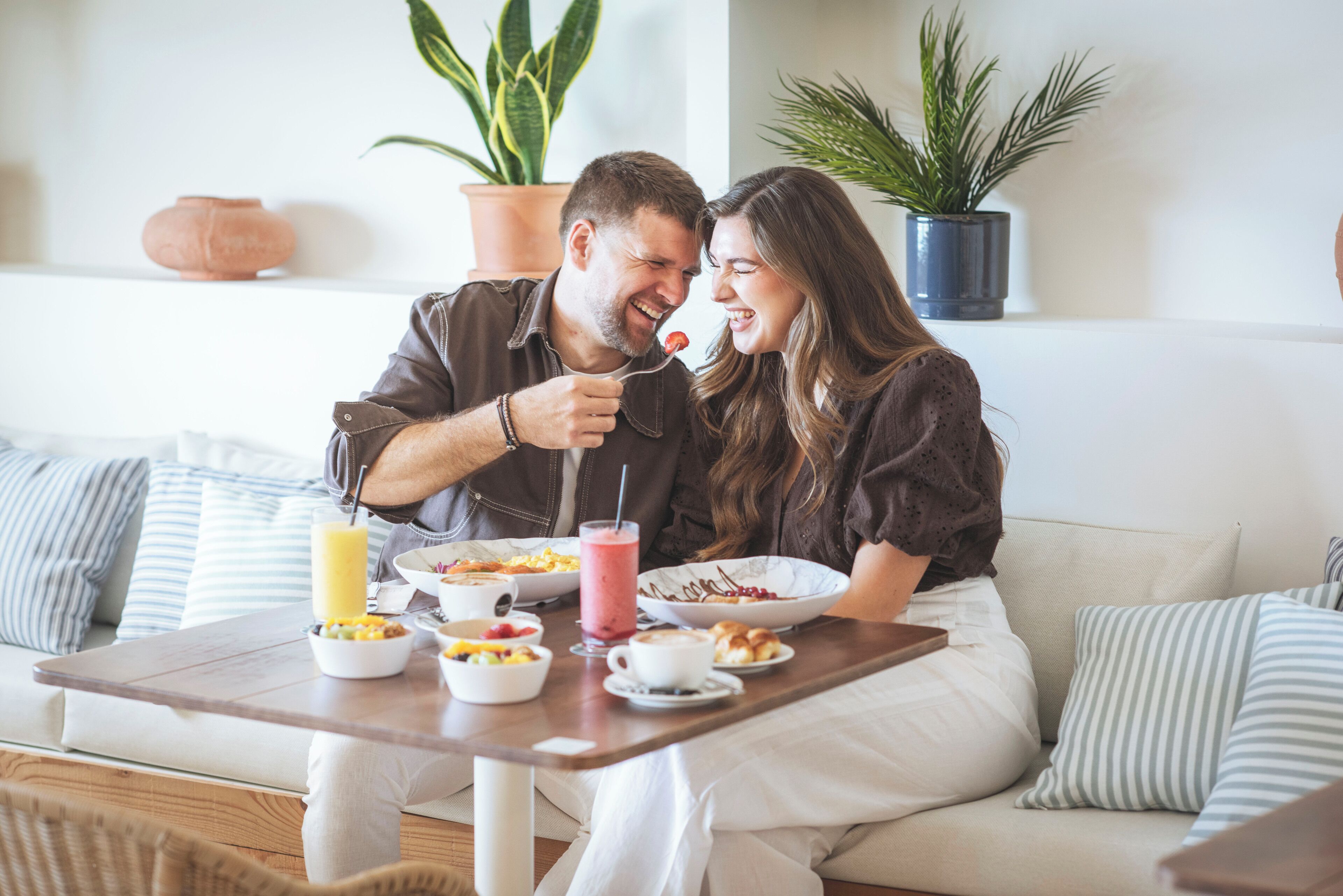 A couple sitting on a sofa with a breakfast table in front of them, featuring bowls of food, a glass of juice, and a coffee cup.