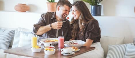 A couple sitting on a sofa with a breakfast table in front of them, featuring bowls of food, a glass of juice, and a coffee cup.