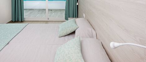 A hotel room with a bed, pillows, a bedside table, a lamp, and a view of the sea through a window with curtains.