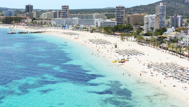 Un paysage urbain côtier avec une vaste plage de sable, des eaux bleues et limpides, et une rangée d’immeubles à plusieurs étages le long de la côte.