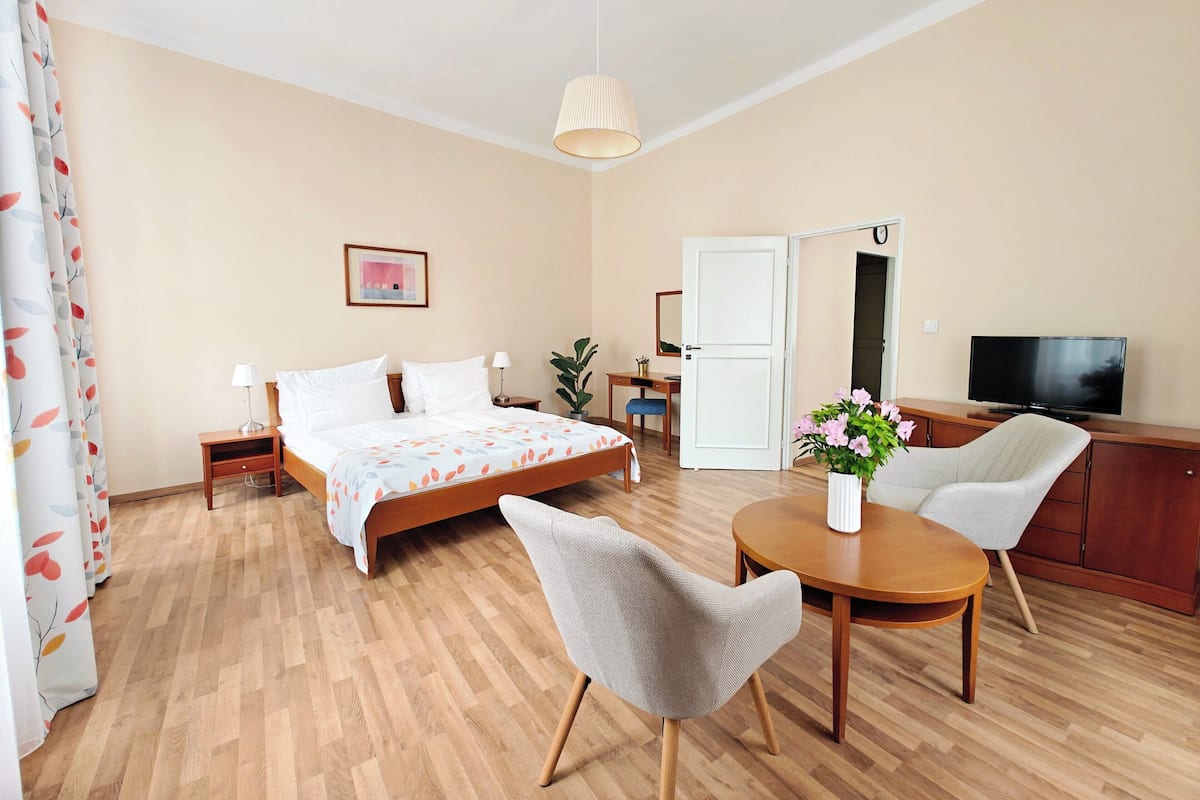 Quarto de hotel com cama, mesas de cabeceira, escrivaninha, cadeira, televisão e mesa de jantar.