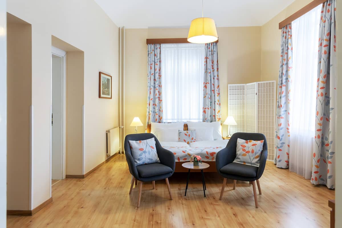 Quarto de hotel com uma cama, duas poltronas, uma mesinha, um abajur e uma janela com cortinas.