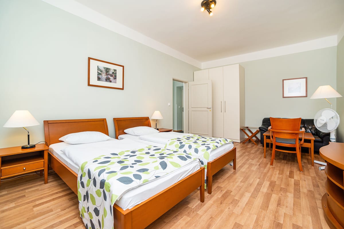 Quarto de hotel com duas camas de solteiro, uma escrivaninha, uma cadeira, um guarda-roupa e um ventilador.