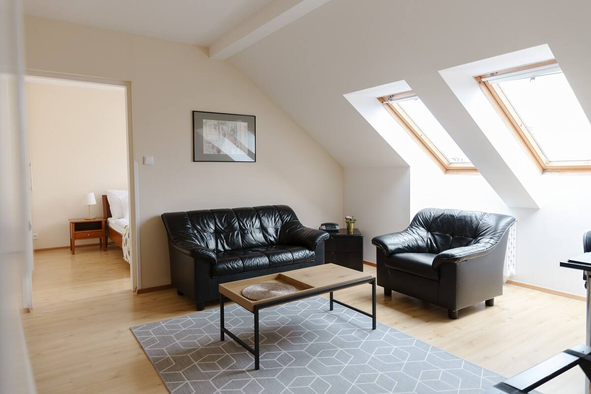 Uma sala de estar moderna com um sofá de couro preto, uma poltrona combinando e uma mesa de centro de madeira. Há claraboias no teto inclinado, um quadro na parede e uma pequena mesa lateral com um abajur.