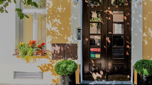 Ein Hotelzugang mit einer dunklen Holztür, je einer von zwei Kübeln mit Pflanzen auf jeder Seite und Blumenkästen an den Fensterbänken.
