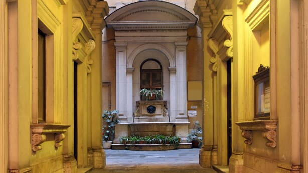 Un pasillo de arquitectura clásica, con columnas y una fuente central adornada con plantas.