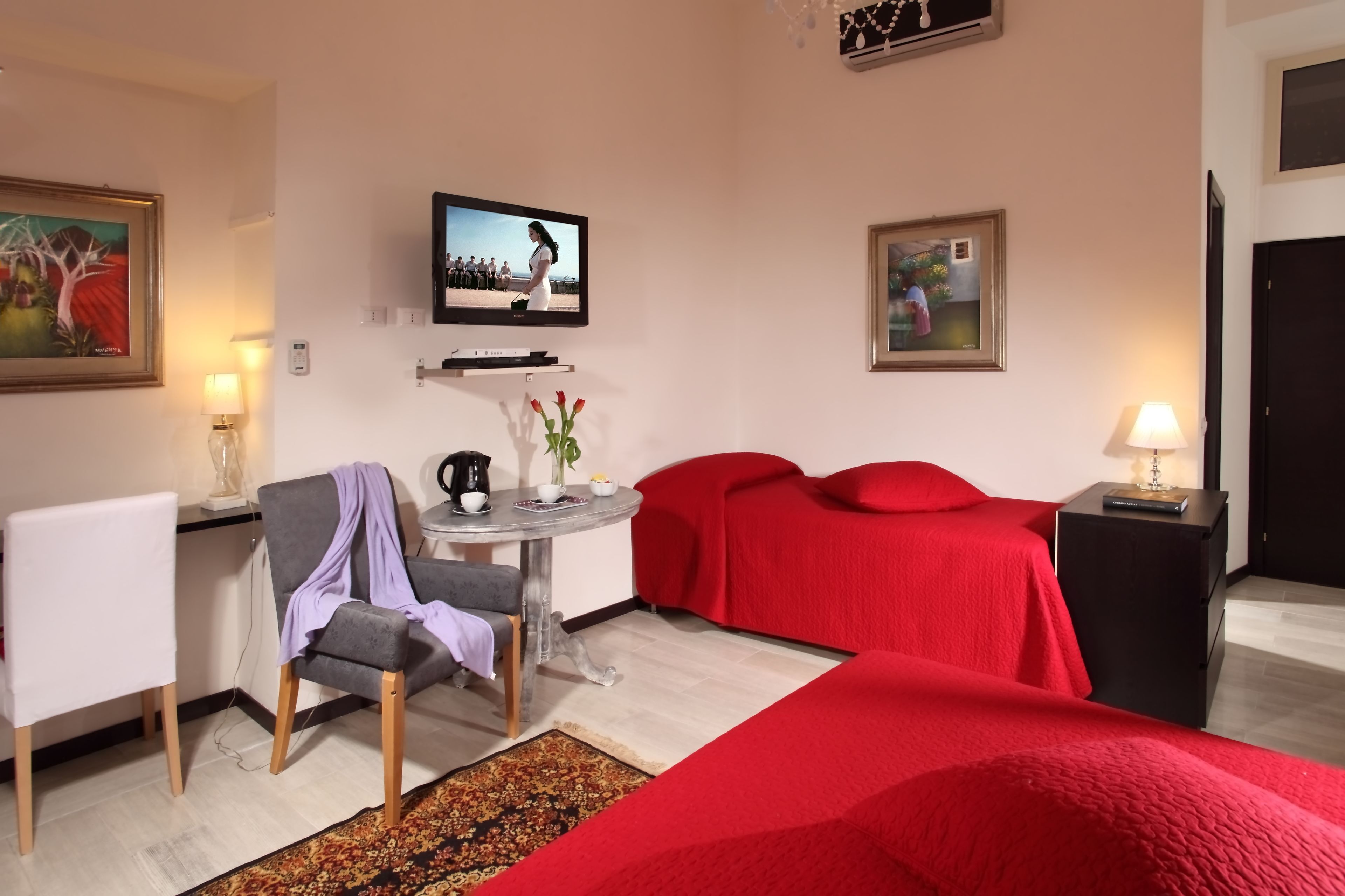 A hotel room with a bed, a desk with a chair, a TV mounted on the wall, and a small table with a vase of flowers.