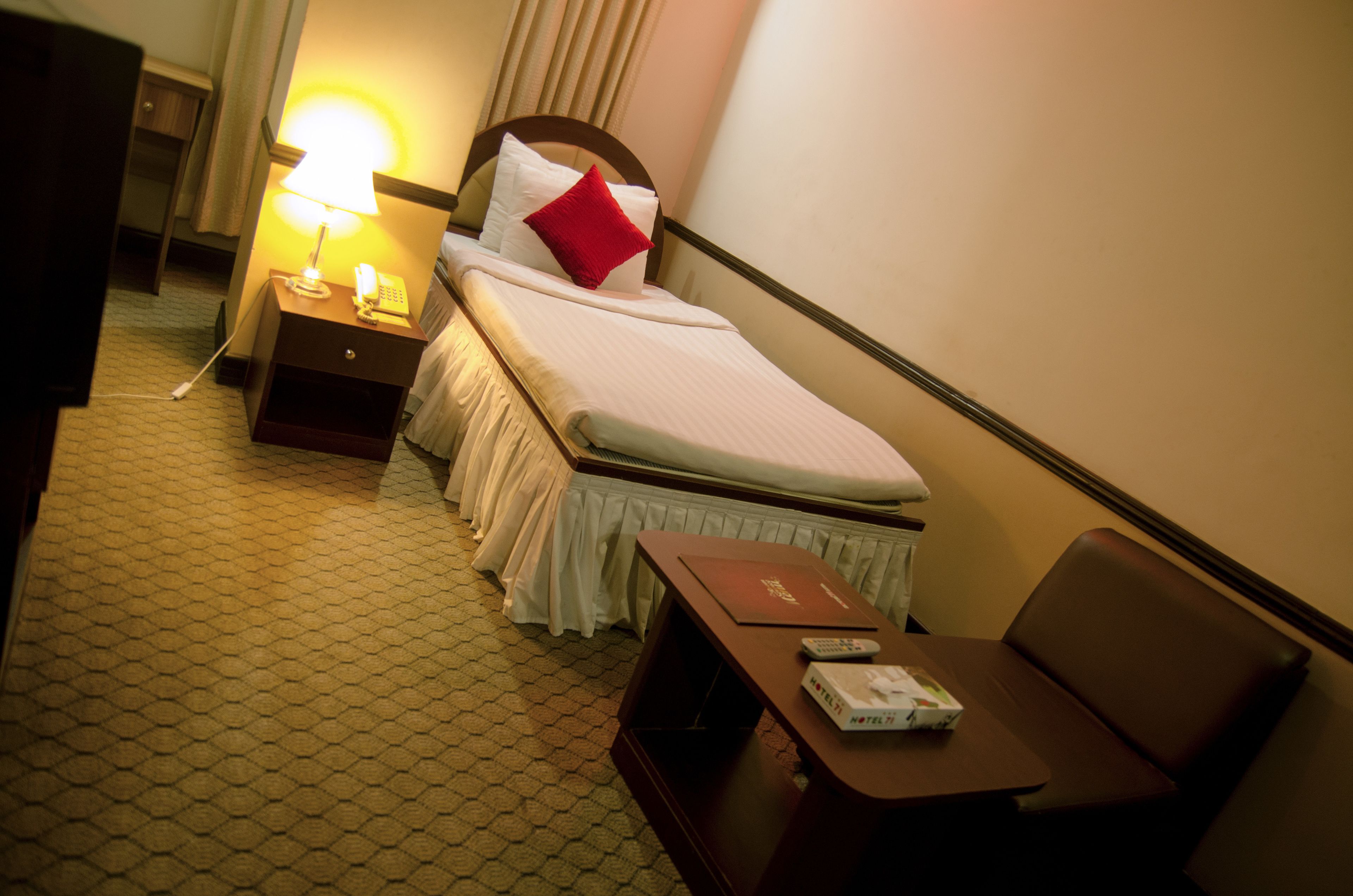 Una habitación de hotel con cama, mesita de noche, lámpara y una pequeña mesa con control remoto y un libro.