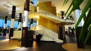 Un moderno vestíbulo de hotel con una escalera central, pilares grandes y plantas en macetas.