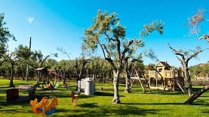 Un parque infantil con columpios y estructuras para escalar, rodeado de árboles y una casa al fondo.