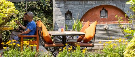 A man seated at a table with orange cushions, a stone structure with a statue, and potted plants in the foreground.