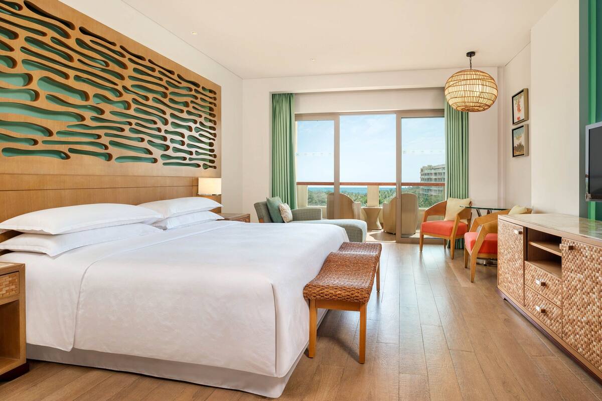 Hotelkamer met een groot bed, een houten hoofdeinde, een geweven bankje, een groen gestreept gordijn en uitzicht op de stad door grote ramen.