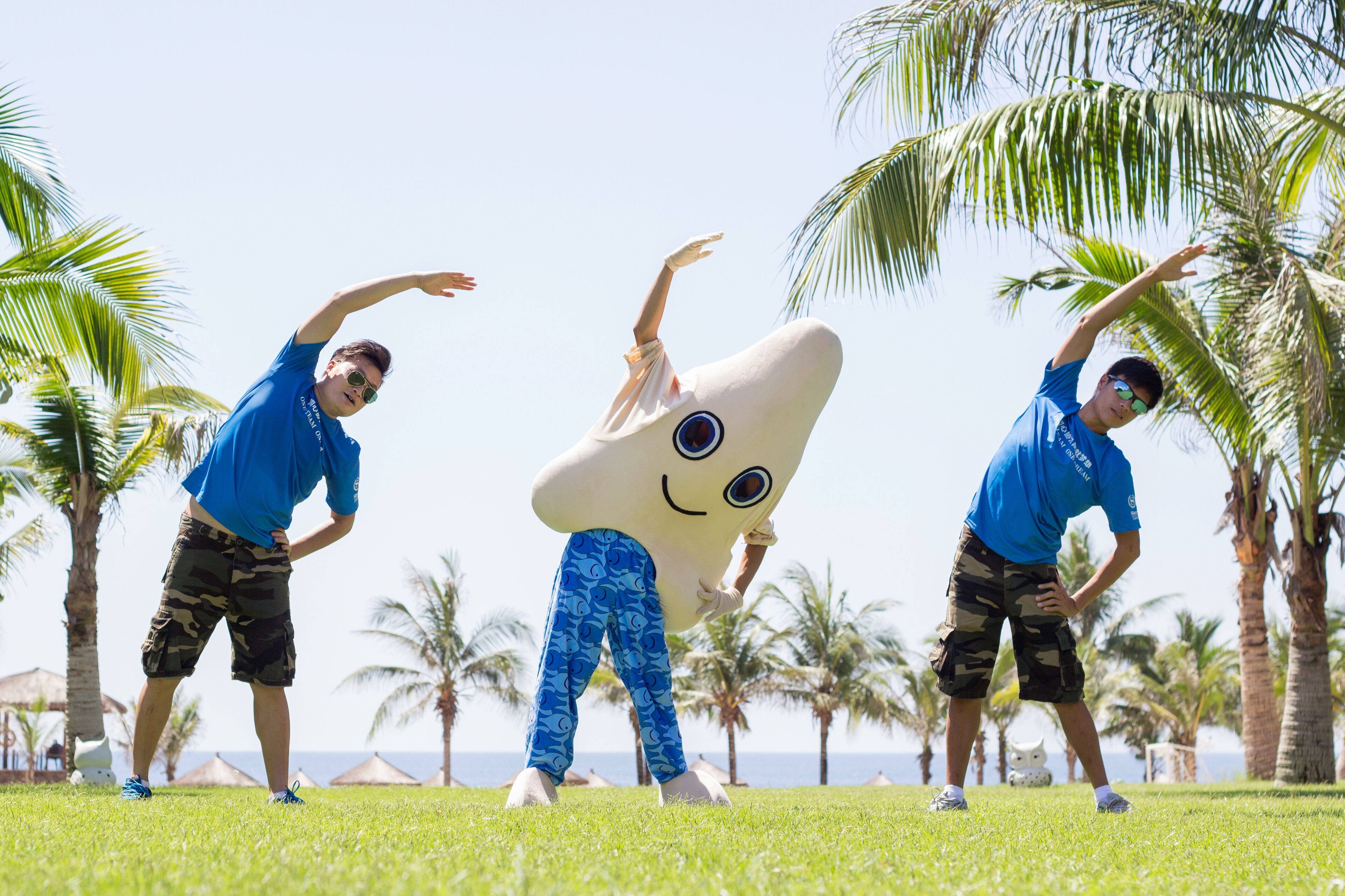 A mascot character in a grassy outdoor area with palm trees and a clear sky.