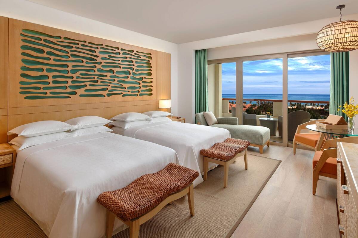Een hotelkamer met twee bedden, een bureau, een stoel, uitzicht op de oceaan en een decoratief paneel aan de muur.