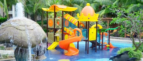 A water play area with a slide and splash pad, surrounded by palm trees and a building in the background.